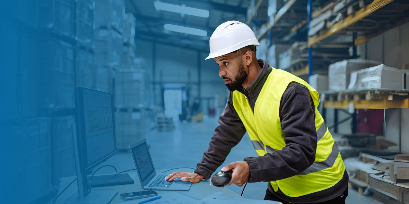 Trabajador de logística con casco y chaleco reflectante, usando un escáner de código de barras para introducir datos en una laptop en el piso de un almacén.