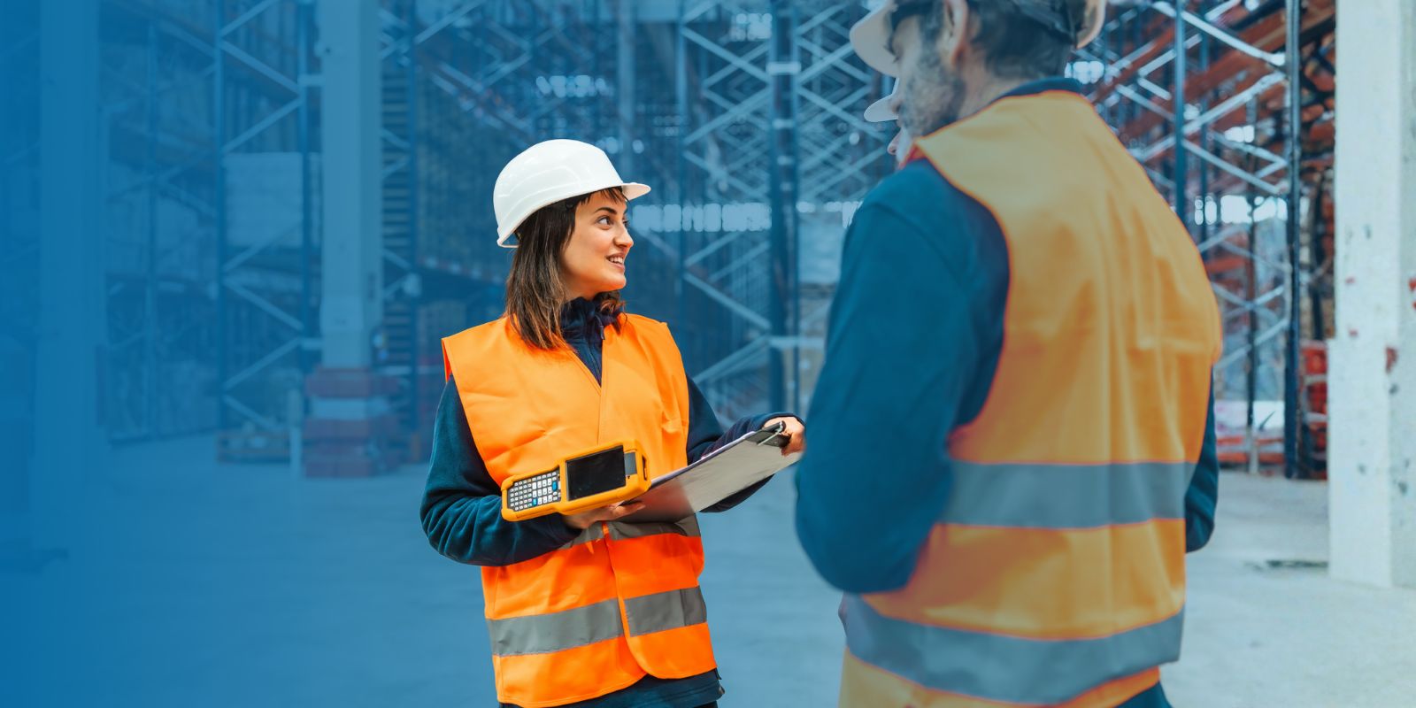 renta-handhelds-honeywell-entrega-inmediata-logistica Ingeniera de logística con casco y chaleco reflectante, coordinando operaciones con un handheld y un portapapeles en un almacén.