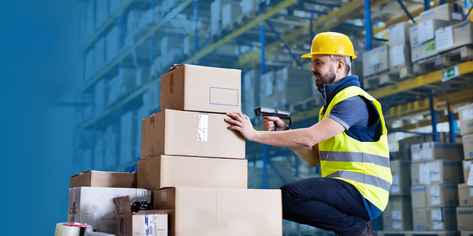 Trabajador de almacén con casco amarillo y chaleco reflectante, usando un dispositivo handheld Honeywell para escanear cajas de cartón en un rack.
