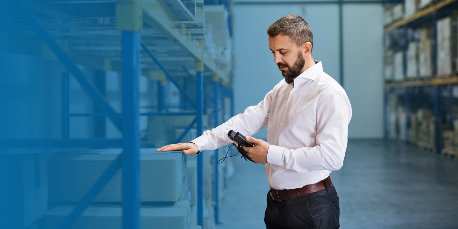 renta-handhelds-zebra-entrega-inmediata-logistica Supervisor o gerente de almacén con camisa blanca, utilizando un escáner handheld para verificar inventario en un pasillo de estanterías industriales.